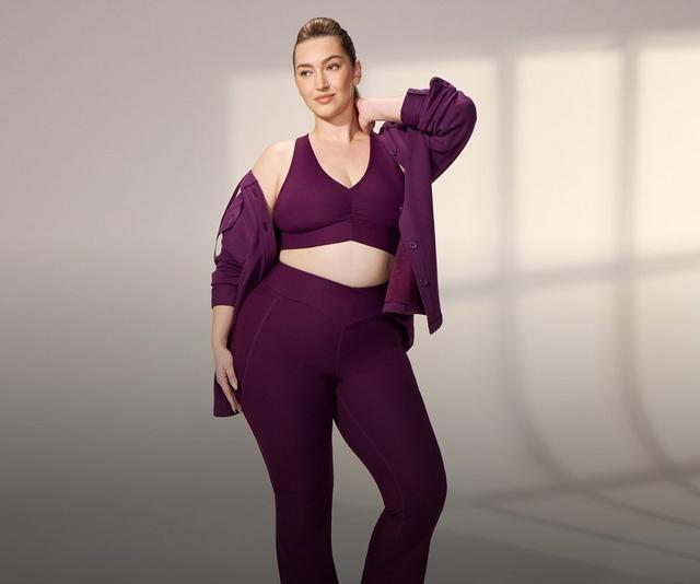 A model with blonde hair pulled back stands in a bright white studio with soft window light on the wall behind her, one hand raised to the back of her neck. She wears a deep plum ribbed sports bra with a V neckline, a ruched center front, and wide shoulder straps, with matching high-waisted ribbed flared leggings that have a V-shaped waistband and seams down the front of the legs. A matching plum button-front shirt jacket hangs off both shoulders behind her, sleeves pushed up, with chest flap pockets and tonal buttons.