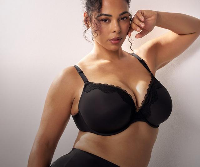 A model with dark curly hair pulled up stands against a soft white wall, one arm bent with her hand near her face and the other at her side. She wears a black bra with molded cups, scalloped lace trim along the upper edge of each cup, a lace-edged center panel, and thin adjustable straps with metal sliders. She also wears black high-waisted briefs with a smooth finish and a plain waistband. Small stud earrings are visible.