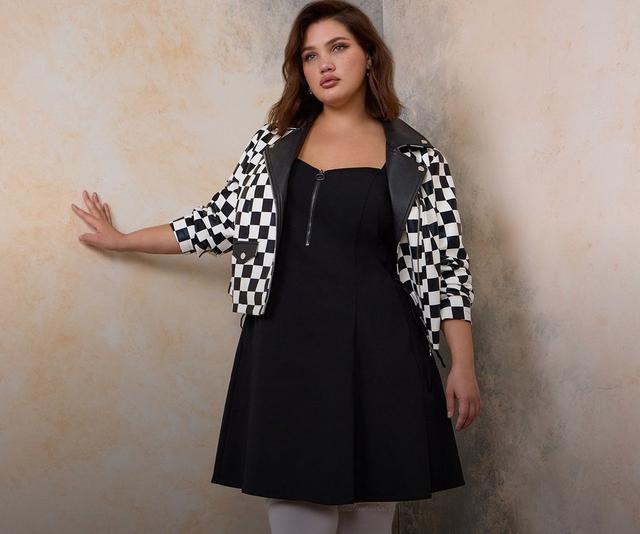 A model with long auburn hair stands in the corner of a room with warm peach mottled walls, grey baseboards, and a herringbone wood floor, one hand pressed flat against the wall beside her. She wears a black and white checkerboard moto jacket with black leather lapels, a zip front, silver hardware, and a flap pocket, open over a sleeveless black fit-and-flare dress with a square neckline and a ring-pull zipper at the chest. She wears opaque white tights and black platform Mary Jane shoes with a T-strap, buckle, and chunky block heel.