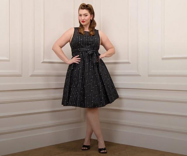 A model with auburn hair styled in vintage victory rolls stands against a cream paneled wall with a herringbone wood floor, both hands on her hips. She wears a sleeveless black fit-and-flare dress with a sheer mesh yoke and boat neckline, printed with fine vertical stripes and scattered silver starbursts, cinched with a matching self-tie bow at the waist and falling into a full pleated knee-length skirt. She wears black patent peep-toe heels, a ring on her right hand, and pearl stud earrings.