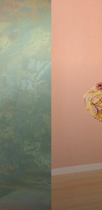 A vertical image split down the middle. The left half shows a mottled sage green and cream painted backdrop with no subject. The right half shows a pale peach plaster wall with white baseboard and light wooden floorboards below. At the right edge, a hand holds out a sweep of yellow fabric printed with pink and burgundy flowers and leafy branches, cropped so only the fabric and fingers are visible.
