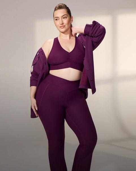 A model with blonde hair pulled back stands in a bright white studio with soft window light on the wall behind her, one hand raised to the back of her neck. She wears a deep plum ribbed sports bra with a V neckline, a ruched center front, and wide shoulder straps, with matching high-waisted ribbed flared leggings that have a V-shaped waistband and seams down the front of the legs. A matching plum button-front shirt jacket hangs off both shoulders behind her, sleeves pushed up, with chest flap pockets and tonal buttons.