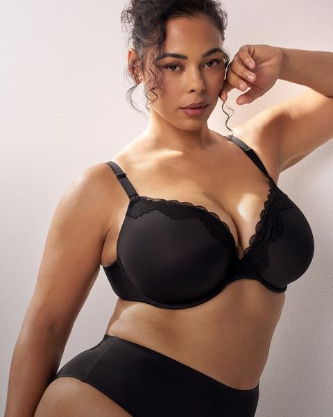 A model with dark curly hair pulled up stands against a soft white wall, one arm bent with her hand near her face and the other at her side. She wears a black bra with molded cups, scalloped lace trim along the upper edge of each cup, a lace-edged center panel, and thin adjustable straps with metal sliders. She also wears black high-waisted briefs with a smooth finish and a plain waistband. Small stud earrings are visible.