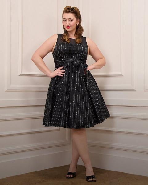 A model with auburn hair styled in vintage victory rolls stands against a cream paneled wall with a herringbone wood floor, both hands on her hips. She wears a sleeveless black fit-and-flare dress with a sheer mesh yoke and boat neckline, printed with fine vertical stripes and scattered silver starbursts, cinched with a matching self-tie bow at the waist and falling into a full pleated knee-length skirt. She wears black patent peep-toe heels, a ring on her right hand, and pearl stud earrings.