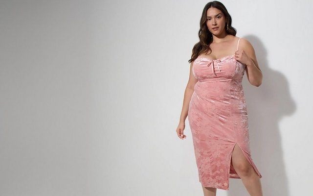 A model with long dark hair poses against a light grey background wearing a pink crushed velvet slip dress with a bow detail and side slit.