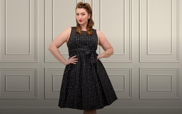 A model with pin-up style wavy brown hair and red lipstick poses against a white paneled wall wearing a blue fit-and-flare dress with a hot air balloon print, sweetheart neckline, wide straps, and a full pleated skirt.