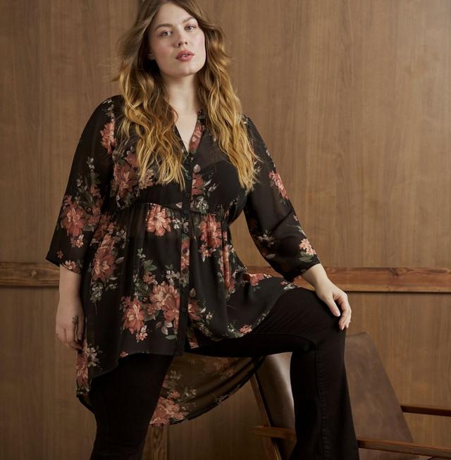 A model with long wavy blonde hair perches on the edge of a wooden bench in front of a warm wood paneled wall, one leg crossed over the other and a hand resting on her knee. She wears a sheer black tunic blouse printed with large coral and dusty rose flowers and sage green leaves, with a V neckline, a full button front, three-quarter bell sleeves, a gathered empire seam, and a high-low hem that sweeps to a long point at the back. Underneath she wears black pants. A small tattoo shows on her left forearm and she wears a thin ring on one hand.