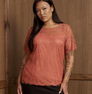 A model with long straight black hair stands in front of a weathered light wood plank wall, arms relaxed at her sides. She wears a coral pink lace top with a scalloped scoop neckline, a sheer illusion yoke across the chest and shoulders, short scalloped sleeves, an allover floral lace pattern, and a scalloped hem, worn over a coral lining. A black skirt or pants shows at the bottom edge. Extensive fine-line tattoos cover both arms.