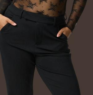 A close-up of a model's hips and upper legs against a dark brown background, both hands tucked into her front pockets. She wears black tailored pants with a flat waistband, belt loops, front pleats, slash side pockets, and a slim straight leg. Above the waistband is a sheer black floral lace bodysuit with long mesh sleeves, visible from the ribcage up.