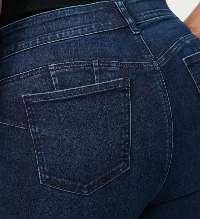 A close-up of a tall stack of folded jeans against a soft blush background. The folded legs face the camera, layering shades of indigo, dark navy, charcoal grey, faded medium blue, and pale light wash. The denim edges curl and fold naturally, showing varied textures, hems, and fading across the stack.