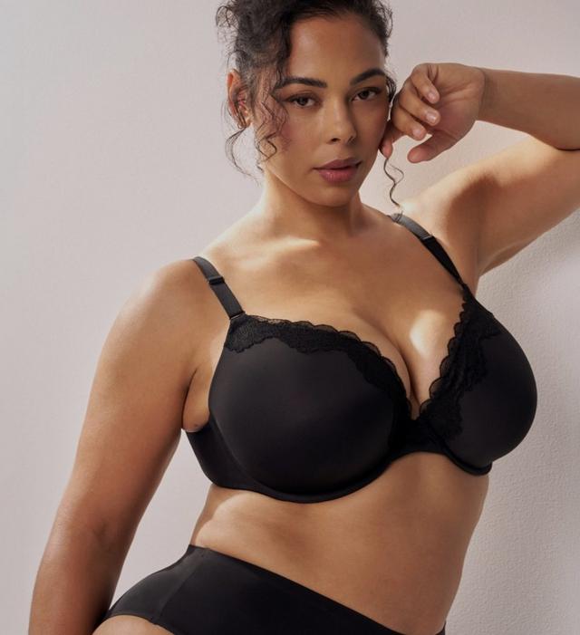 A model with dark curly hair pulled up stands at the right side of a plain soft white wall, one arm raised with her hand near her ear and her body angled toward the camera. She wears a black bra with smooth molded cups, scalloped lace trim along the upper edge of each cup and the center panel, and thin adjustable straps with metal sliders. She also wears black high-waisted briefs with a plain wide waistband. The left portion of the frame is empty wall.