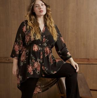 A model with long wavy blonde hair perches on the edge of a wooden bench in front of a warm wood paneled wall, one leg crossed over the other and a hand resting on her knee. She wears a sheer black tunic blouse printed with large coral and dusty rose flowers and sage green leaves, with a V neckline, a full button front, three-quarter bell sleeves, a gathered empire seam, and a high-low hem that sweeps to a long point at the back. Underneath she wears black pants. A small tattoo shows on her left forearm and she wears a thin ring on one hand.