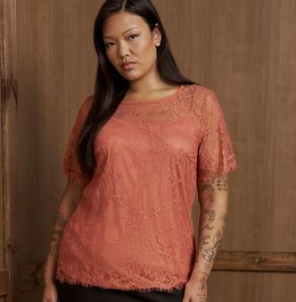 A model with long straight black hair stands in front of a weathered light wood plank wall, arms relaxed at her sides. She wears a coral pink lace top with a scalloped scoop neckline, a sheer illusion yoke across the chest and shoulders, short scalloped sleeves, an allover floral lace pattern, and a scalloped hem, worn over a coral lining. A black skirt or pants shows at the bottom edge. Extensive fine-line tattoos cover both arms.