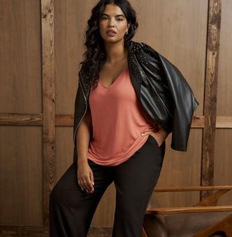 A model with long dark curly hair stands in front of a wood paneled wall, one hand in her pocket, with a black faux leather moto jacket draped over both shoulders. The jacket has silver zippers and clusters of gold studs across the shoulders. Underneath she wears a coral pink V-neck top with a relaxed drape, tucked loosely at the front, and black wide-leg trousers. She wears gold hoop earrings and rings on one hand.