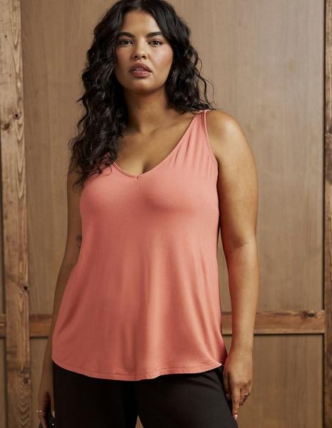 A model with long dark curly hair stands facing the camera in front of a weathered light wood plank wall with vertical boards and visible grain. She wears a coral pink knit tank top with a V neckline, wide shoulder straps, a relaxed swing silhouette, and a curved hem. Black pants are visible at the bottom of the frame. She wears small gold hoop earrings, a ring on each hand, and a small tattoo shows on her inner forearm.