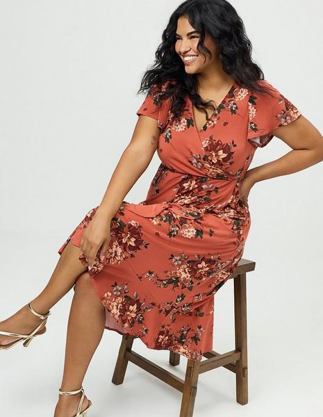 A model with long dark curly hair sits on a wooden stool against a plain white background, smiling and looking off to her right with one hand behind her back. She wears a rust orange midi dress printed with burgundy, peach, and cream florals and dark green leaves, with a surplice V neckline, flutter cap sleeves, a twisted knot detail at the waist, and a ruffled wrap skirt with a high front slit. On her feet are gold strappy heeled sandals with ankle straps, and a small tattoo is visible on her upper arm.
