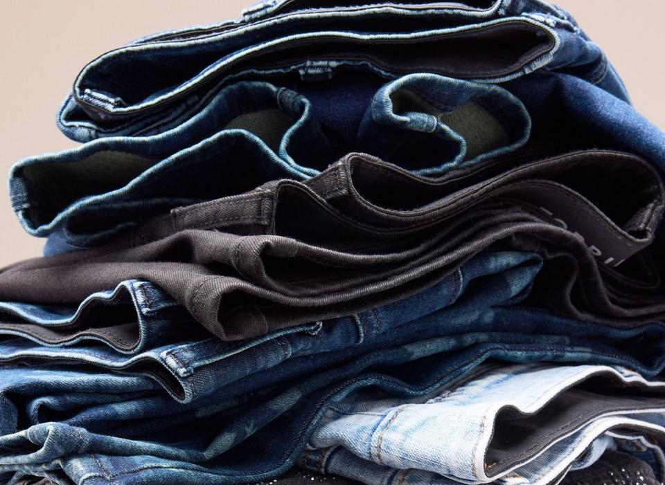 A close-up back view of a model wearing dark-wash blue jeans, cropped from the waistband to the upper thigh. One back pocket fills the center of the frame, edged with topstitching in a matching thread and set below a curved yoke seam. The denim shows subtle fading and whiskering across the seat, with belt loops and the waistband visible along the top edge. A section of bare skin and the model's other leg appear at the right edge against a plain light background.