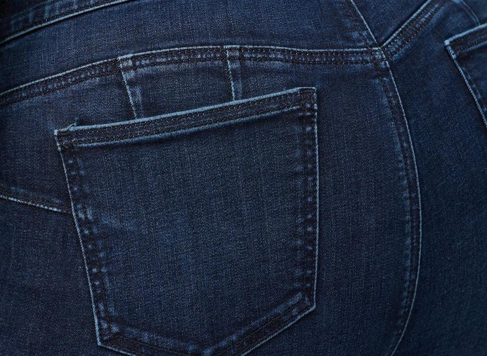 A close-up of a tall stack of folded jeans against a soft blush background. The folded legs face the camera, layering shades of indigo, dark navy, charcoal grey, faded medium blue, and pale light wash. The denim edges curl and fold naturally, showing varied textures, hems, and fading across the stack.