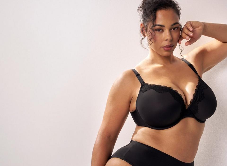 A model with dark curly hair pulled up stands at the right side of a plain soft white wall, one arm raised with her hand near her ear and her body angled toward the camera. She wears a black bra with smooth molded cups, scalloped lace trim along the upper edge of each cup and the center panel, and thin adjustable straps with metal sliders. She also wears black high-waisted briefs with a plain wide waistband. The left portion of the frame is empty wall.