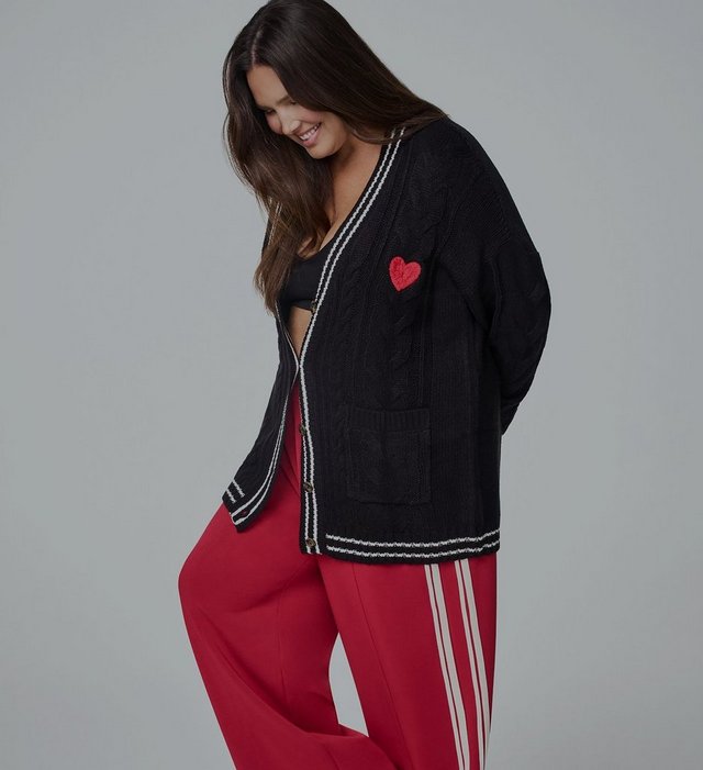 A close-up of a model with long dark wavy hair from the head to the waist against a plain light grey background, looking directly at the camera. She wears a fitted red ribbed knit henley with long sleeves, a scooped neckline trimmed in contrasting white, and a white button placket down the center chest fastened with four gold-tone buttons. A white stripe detail shows on the cuff at the lower right, and black pants appear at the bottom edge.