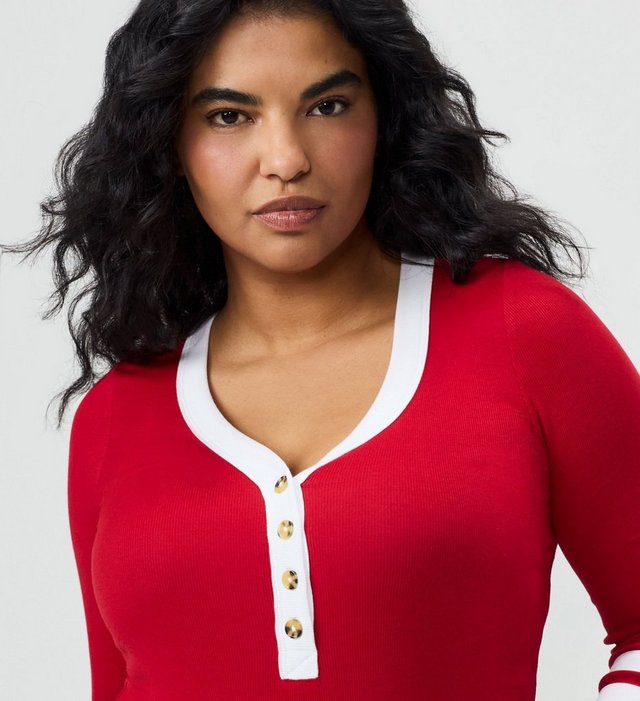 A close-up of a model with long dark wavy hair from the head to the waist against a plain light grey background, looking directly at the camera. She wears a fitted red ribbed knit henley with long sleeves, a scooped neckline trimmed in contrasting white, and a white button placket down the center chest fastened with four gold-tone buttons. A white stripe detail shows on the cuff at the lower right, and black pants appear at the bottom edge.