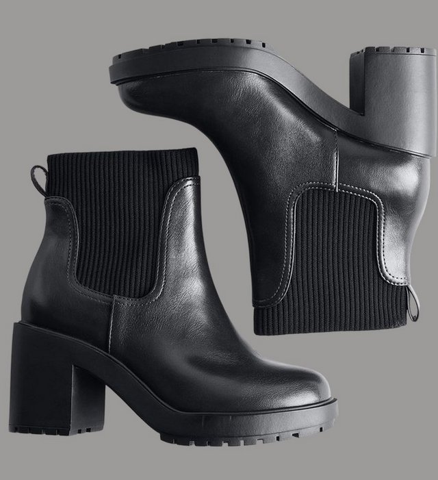 A pair of black ankle boots arranged on a grey background, one standing upright and the other lying on its side above it. The upright boot has a smooth black leather shaft with a ribbed knit panel at the back, a pull tab at the heel, a rounded toe, a chunky lugged platform sole, and a tall block heel. The boot on its side shows the underside of the treaded sole and the ribbed knit collar with its metal pull loop.