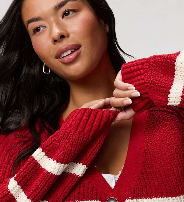 A close-up of a model with long dark hair, looking off to her left with both hands raised near her chin so her sleeves fall back. She wears a chunky red knit cardigan with wide cream horizontal stripes, a ribbed V neckline, dropped shoulders, deep ribbed cuffs, and a dark button at the center front. A white top with a button and lace trim shows at the neckline. She wears a gold rectangular hoop earring and has white French-tip nails.