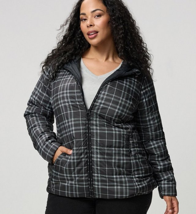 A model with long dark curly hair stands facing the camera against a plain light grey background, one hand tucked into a pocket. She wears a black and white plaid puffer jacket with a glossy finish, a full zip front with a ring pull, a stand collar, quilted horizontal panels, and side pockets. A grey V-neck top shows underneath, and she wears black jeans.