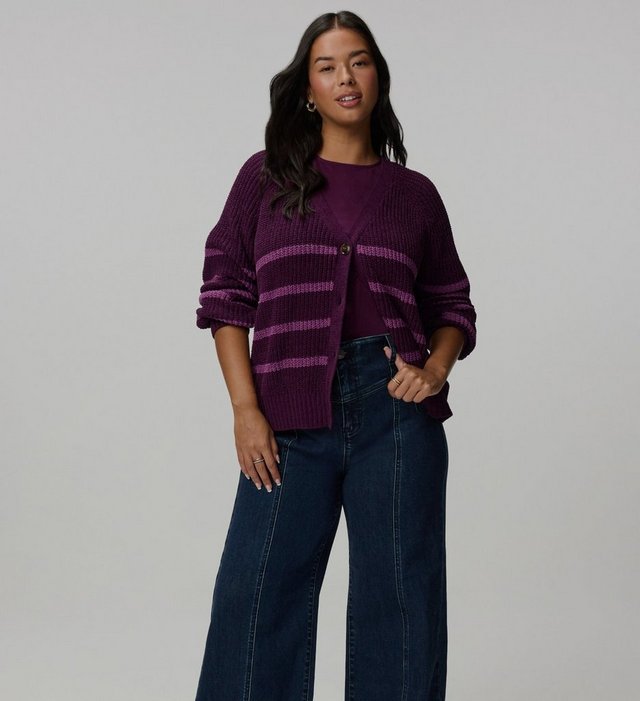 A model with long dark wavy hair stands facing the camera against a plain light grey background, arms relaxed at her sides. She wears a deep plum chunky knit cardigan with lighter purple horizontal stripes across the body and sleeves, a V neckline, dropped raglan shoulders, pushed-up sleeves, and a single tortoiseshell button at the chest. A matching plum crewneck top shows underneath. Her jeans are dark-wash and wide-legged with a seam down the front of each leg and a curved yoke at the hip. She wears small gold hoop earrings and rings on both hands.
