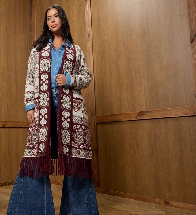 A model with long dark wavy hair stands at the left side of a warm wood paneled wall, looking off to her right with one hand raised to the front of her cardigan. She wears a long open duster cardigan knit in an allover folk-style floral pattern of cream, burgundy, olive green, and brown medallions, with a wide burgundy patterned border down the front placket, patch pockets at the hip, and fringe along the hem. Underneath she wears a light blue chambray button-down shirt with the sleeves rolled and dark-wash jeans. She wears small hoop earrings, rings on both hands, and pale manicured nails. The right half of the frame is empty wood paneling.