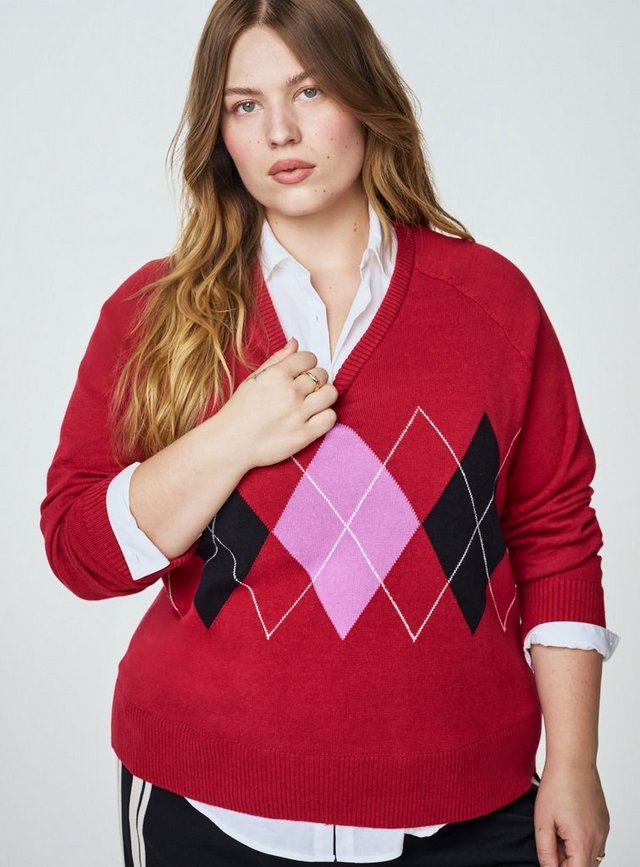 A model with long wavy strawberry-blonde hair is shown from the head to the hips against a plain light grey background, one hand raised to the neckline of her sweater. She wears a red V-neck knit sweater with a large argyle pattern across the lower front in pink, black, and white diamond shapes with thin white crosshatch lines, finished with ribbed cuffs, neckline, and hem. A crisp white collared shirt is layered underneath, its collar, cuffs, and hem showing. Black pants with a cream side stripe appear at the bottom edge, and she wears a gold ring.
