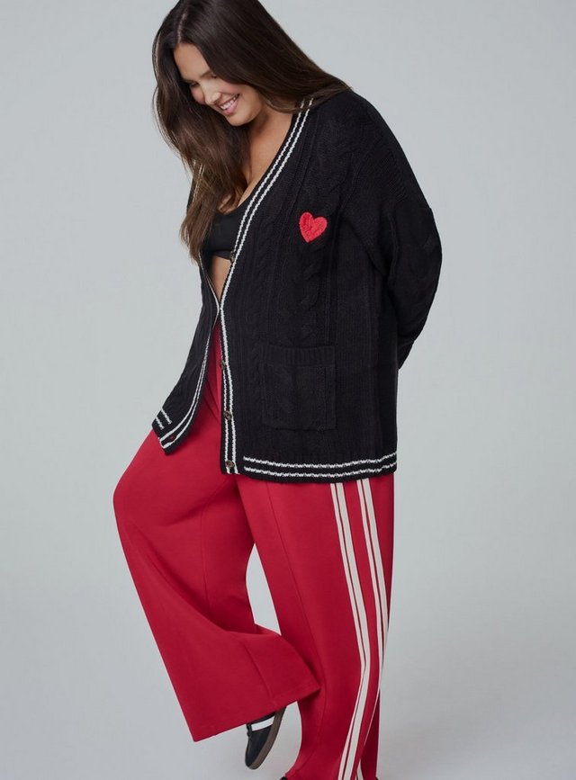 A close-up of a model with long dark wavy hair from the head to the waist against a plain light grey background, looking directly at the camera. She wears a fitted red ribbed knit henley with long sleeves, a scooped neckline trimmed in contrasting white, and a white button placket down the center chest fastened with four gold-tone buttons. A white stripe detail shows on the cuff at the lower right, and black pants appear at the bottom edge.