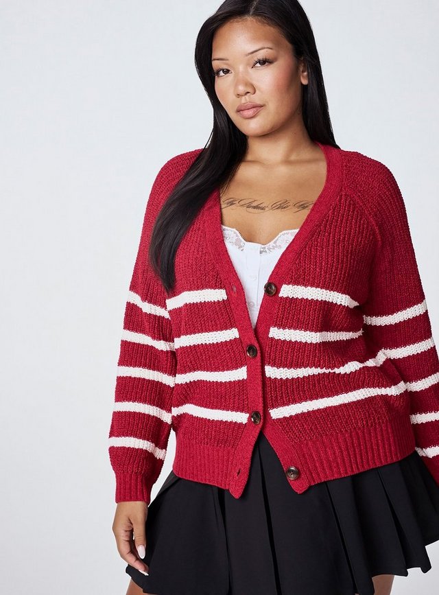 A model with long straight black hair is shown from the head to the hips against a plain light grey background. She wears a red chunky knit cardigan with cream horizontal stripes, a deep V neckline, dropped raglan shoulders, ribbed cuffs and hem, and tortoiseshell buttons down the front. Underneath is a white top with lace trim and a button placket. She wears a black pleated skirt, has white nails, and has script lettering tattooed across her upper chest.