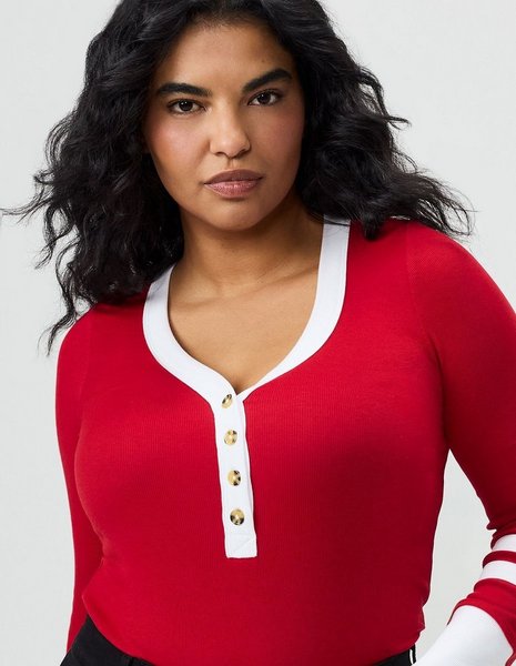A close-up of a model with long dark wavy hair from the head to the waist against a plain light grey background, looking directly at the camera. She wears a fitted red ribbed knit henley with long sleeves, a scooped neckline trimmed in contrasting white, and a white button placket down the center chest fastened with four gold-tone buttons. A white stripe detail shows on the cuff at the lower right, and black pants appear at the bottom edge.