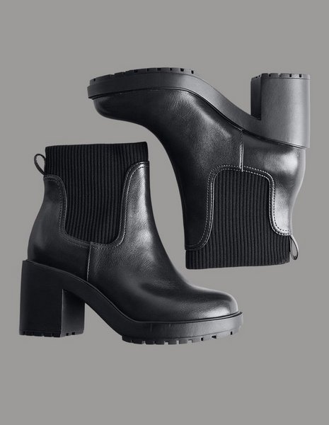 A pair of black ankle boots arranged on a grey background, one standing upright and the other lying on its side above it. The upright boot has a smooth black leather shaft with a ribbed knit panel at the back, a pull tab at the heel, a rounded toe, a chunky lugged platform sole, and a tall block heel. The boot on its side shows the underside of the treaded sole and the ribbed knit collar with its metal pull loop.