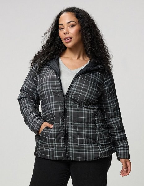 A model with long dark curly hair stands facing the camera against a plain light grey background, one hand tucked into a pocket. She wears a black and white plaid puffer jacket with a glossy finish, a full zip front with a ring pull, a stand collar, quilted horizontal panels, and side pockets. A grey V-neck top shows underneath, and she wears black jeans.