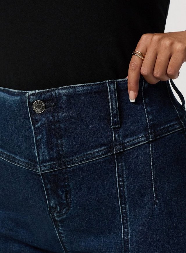 A close-up of a model's waist and hip, with one hand hooked into the waistband of her jeans. The dark-wash denim shows a silver shank button stamped with 'TORRID' in a circle, a belt loop, a curved yoke seam across the front, a seam running down the leg, and a small coin pocket. Light fading runs along the seams and creases. She wears a black top tucked above the waistband, a thin gold ring, and white French-tip nails.