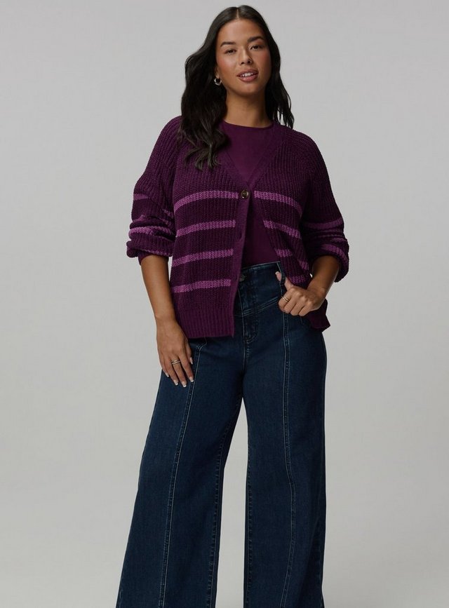 A model with long dark wavy hair stands facing the camera against a plain light grey background, arms relaxed at her sides. She wears a deep plum chunky knit cardigan with lighter purple horizontal stripes across the body and sleeves, a V neckline, dropped raglan shoulders, pushed-up sleeves, and a single tortoiseshell button at the chest. A matching plum crewneck top shows underneath. Her jeans are dark-wash and wide-legged with a seam down the front of each leg and a curved yoke at the hip. She wears small gold hoop earrings and rings on both hands.