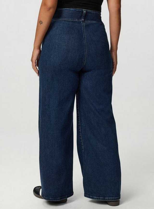 A back view of a model from the waist down against a plain light grey background. She wears dark-wash wide-leg jeans with a wide smooth waistband, a horizontal yoke seam across the back, a center back seam, and no back pockets, falling in a straight wide line to the floor. Black and gum-soled sneakers show beneath the hems. A black top and small tattoos on both arms are visible at the top edge.