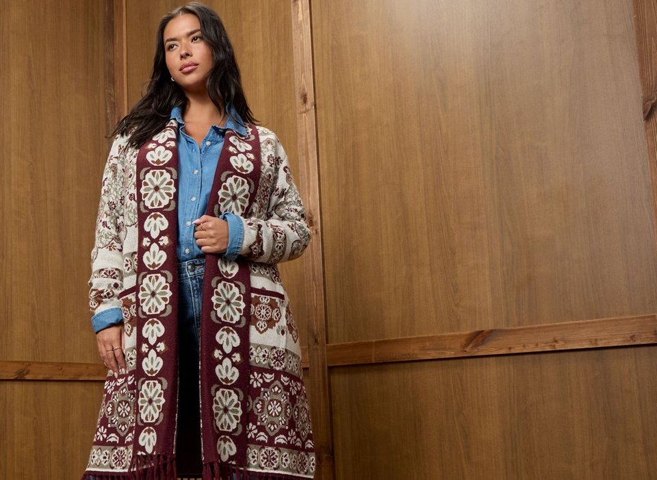 A model with long dark wavy hair stands at the left side of a warm wood paneled wall, looking off to her right with one hand raised to the front of her cardigan. She wears a long open duster cardigan knit in an allover folk-style floral pattern of cream, burgundy, olive green, and brown medallions, with a wide burgundy patterned border down the front placket, patch pockets at the hip, and fringe along the hem. Underneath she wears a light blue chambray button-down shirt with the sleeves rolled and dark-wash jeans. She wears small hoop earrings, rings on both hands, and pale manicured nails. The right half of the frame is empty wood paneling.