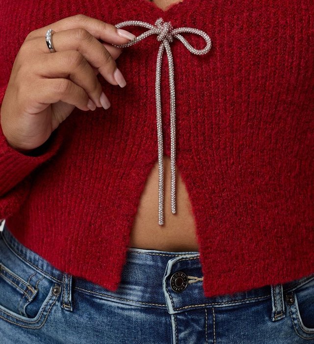 A close-up of a model's torso against a plain background, showing a cropped red knit cardigan with a deep V opening at the center front and a silver metallic drawstring tied in a long bow at the chest. One hand is raised to hold the end of the bow, with a silver ring on the finger. Light-wash blue jeans with a button closure show below the cropped hem, and a strip of midriff is visible.