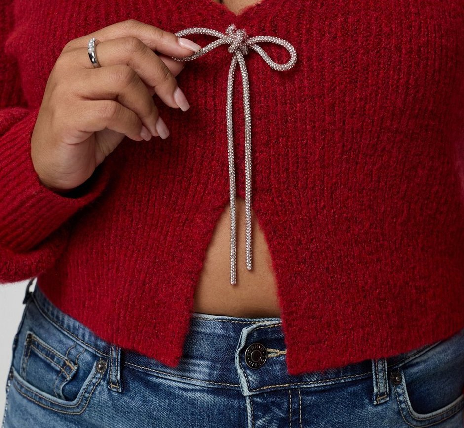 A close-up of a model's torso against a plain background, showing a cropped red knit cardigan with a deep V opening at the center front and a silver metallic drawstring tied in a long bow at the chest. One hand is raised to hold the end of the bow, with a silver ring on the finger. Light-wash blue jeans with a button closure show below the cropped hem, and a strip of midriff is visible.