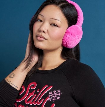 A close-up of a model with long black hair against a deep blue background, one hand raised to the side of her neck. She wears bright pink fluffy earmuffs with a padded headband, and a black sweatshirt printed in red script and white snowflakes, with the partial word 'Still' visible across the chest. A small tattoo shows on her forearm.