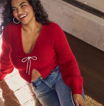 A model with dark curly hair laughs while leaning back, shown from the head to the hips in a warm indoor setting with a stone fireplace and a textured throw behind her. She wears a fuzzy red knit cardigan with a deep V opening, long sleeves, and a metallic drawstring tied in a bow at the chest, cropped to show a sliver of midriff. She wears light-wash blue jeans and large gold hoop earrings.