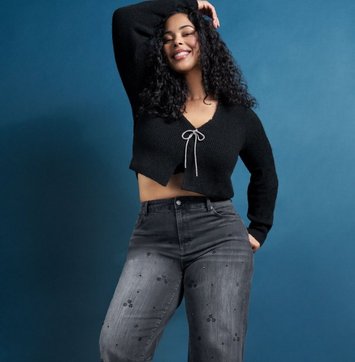 A model with dark curly hair smiles with one arm raised overhead against a deep blue background. She wears a cropped black knit cardigan with long sleeves, a V opening, and a silver metallic drawstring tied in a bow at the chest. Her jeans are washed grey with small scattered embellishments down the leg.