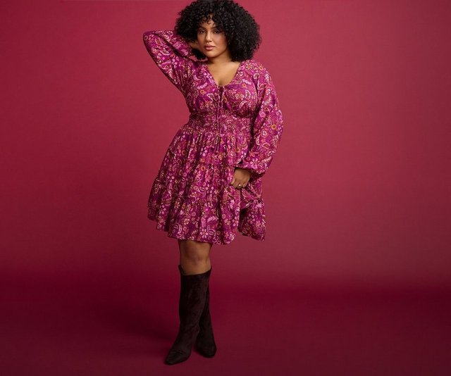 A model with dark curly hair stands against a solid deep burgundy background, one hand raised toward her head and the other at her side. She wears a short magenta and burgundy floral-print dress with a V neckline, long blouson sleeves gathered at the cuffs, a fitted bodice with a seamed waist, and a tiered flared skirt ending above the knee. On her feet are tall dark brown boots that reach just below the knee.