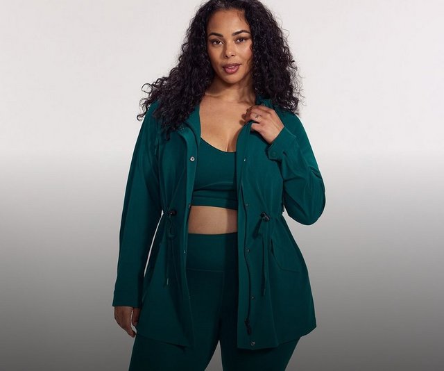 A model with long dark curly hair stands against a plain pale grey background, one hand raised to hold the open front of her jacket. She wears a deep teal green anorak-style jacket with a hood, a zip front with a snap-button storm placket, long sleeves with buttoned cuffs, flap pockets at the hip, and drawstring ties with toggles at the waist and hem. Underneath she wears a matching teal sports bra with a V neckline and a wide underband, leaving her midriff bare, and matching high-waisted teal leggings. She wears a ring on her right hand.