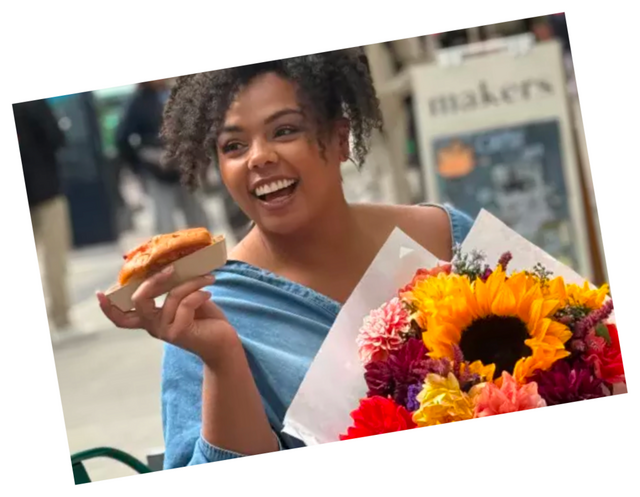 A photo tilted at a slight angle with a thin white border, set against a plain white background. A woman with curly dark hair laughs with her head tilted back, holding a pastry in one hand and a paper-wrapped bouquet in the other. The bouquet contains a large sunflower along with orange, red, pink, and yellow blooms. She wears a light blue denim jacket or shirt off one shoulder over a dark top. Behind her is a blurred street scene with a storefront sign reading ‘makers’, a bench, and greenery.