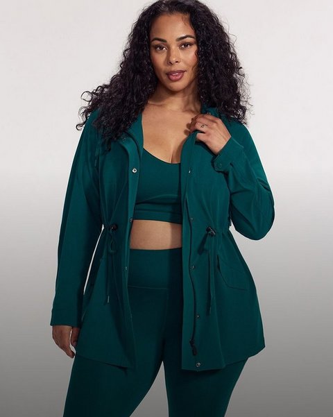 A model with long dark curly hair stands against a plain pale grey background, one hand raised to hold the open front of her jacket. She wears a deep teal green anorak-style jacket with a hood, a zip front with a snap-button storm placket, long sleeves with buttoned cuffs, flap pockets at the hip, and drawstring ties with toggles at the waist and hem. Underneath she wears a matching teal sports bra with a V neckline and a wide underband, leaving her midriff bare, and matching high-waisted teal leggings. She wears a ring on her right hand.