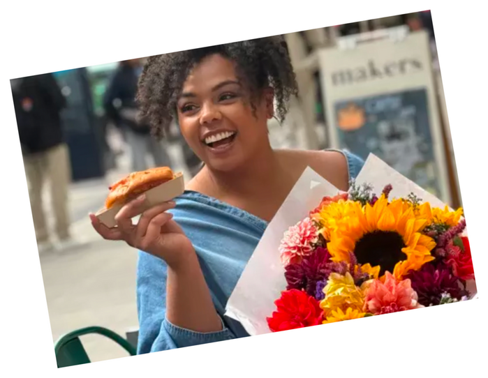 A photo tilted at a slight angle with a thin white border, set against a plain white background. A woman with curly dark hair laughs with her head tilted back, holding a pastry in one hand and a paper-wrapped bouquet in the other. The bouquet contains a large sunflower along with orange, red, pink, and yellow blooms. She wears a light blue denim jacket or shirt off one shoulder over a dark top. Behind her is a blurred street scene with a storefront sign reading ‘makers’, a bench, and greenery.